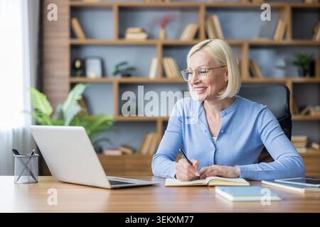 Mature businesswoman wearing glasses and a blue cardigan actively engaging in work, learning, or remote education at her home office, smiling and taking notes while looking at her laptop screen Stock Photo