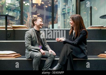 Happy male and female business professionals talking with each other while sitting on steps during networking event in c Stock Photo