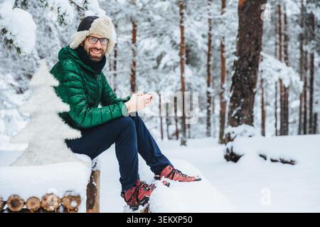 Photo of cheerful male types messages on smart phone looks directly into camera, sits in white winter forest, smiles happily, enjoys communication, watches video, admires landscapes Stock Photo