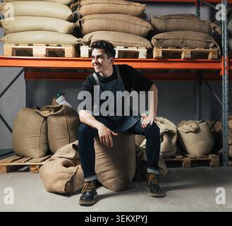 Cheerful male warehouse worker sitting on a large coffee bean bag inside a storage facility. The worker is wearing a protective apron, reinforcing his role in handling goods and participating in daily warehouse operations. He is captured in a relaxed seated position, looking away from the camera with a satisfied expression, during a busy shift. Stock Photo