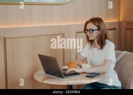 Image of happy smiling woman in earphones using laptop while sitting at cafe, young female freelancer working remotely on laptop computer in coffee shop, studying online during morning breakfast Stock Photo