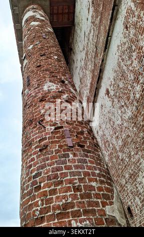 The old red brick column stands tall, showing signs of weathering and age. It contrasts against the worn wall of a taller building nearby. Both struct Stock Photo
