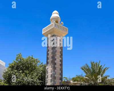 A tall, cylindrical clock tower with a white top stands prominently in an urban park, surrounded by green trees under a bright blue sky. Stock Photo
