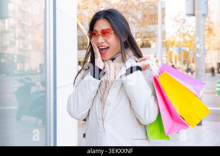 Surprised Asian woman with heart glasses holding colorful shopping bags on the street Stock Photo