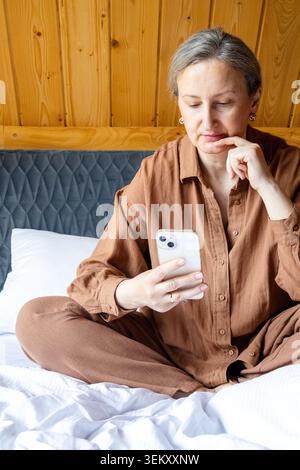Thoughtful middle aged woman using smartphone while sitting on bed, making a decision and comparing options online. Modern lifestyle and choice concep Stock Photo