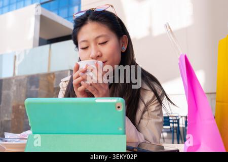 Asian woman holding a coffee cup and using a tablet in a modern outdoor cafe area Stock Photo