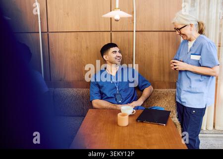 Smiling senior female nurse talking with male coworker sitting on sofa during coffee break in hospital cafeteria Stock Photo