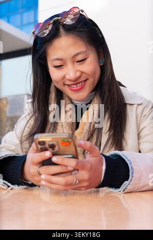 Asian woman using a smartphone in a modern outdoor cafe with colorful shopping bags Stock Photo