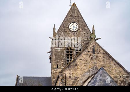 A close-up view of the monumental stone bell tower of Sainte-Mère-Église church, Normandy. The photo captures the clock face on the gable, medieval wi Stock Photo