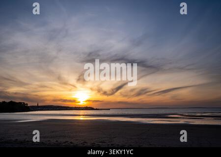 Scenic view of the low sun over the calm sea in Douarnenez. Golden light reflects on the wet sandy shore, while dramatic dark clouds fan out across th Stock Photo