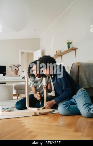 Boy and girl helping each other assembling wooden chair while sitting in living room at home Stock Photo