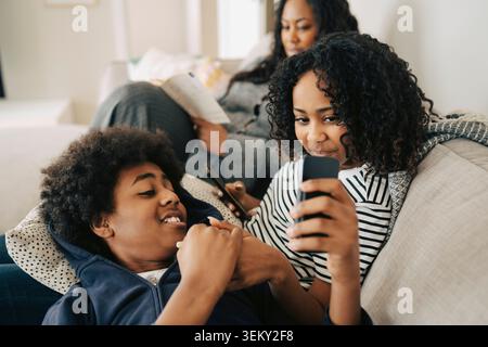 Boy sharing smart phone with sister sitting on sofa with family at home Stock Photo
