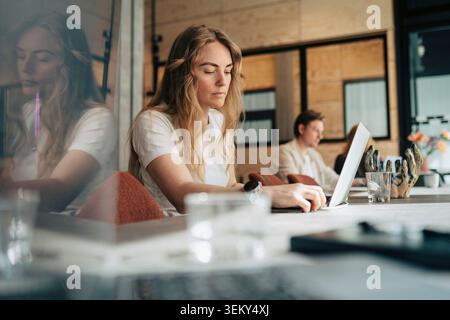 Businesswoman connecting cable into laptop while sitting at shared desk in coworking hub Stock Photo