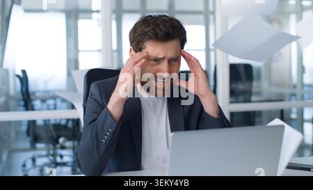 Stressed Caucasian mature business man feeling headache looking in laptop flying papers documents around overload middle-aged businessman male look in Stock Photo