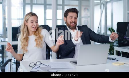 Two business professionals using laptop typing computer rejoicing dancing work complete job done at office table happy Caucasian businessman with Stock Photo