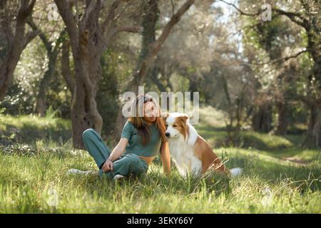 A woman gently hugs her Border Collie while sitting in a sun-dappled forest. Their bond is visible in this peaceful, emotional moment. Stock Photo