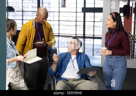 Diverse coworkers collaborating by windows in office, seated man in wheelchair holding tablet Stock Photo