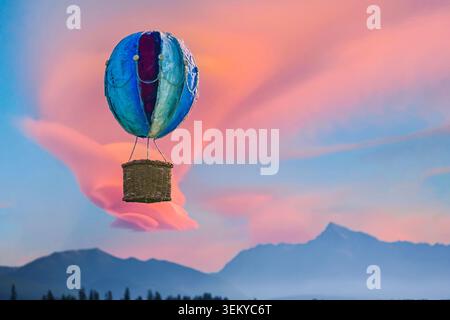 Hot air balloon floating above mountains with pink clouds forming a hand shape in the sky. Stock Photo
