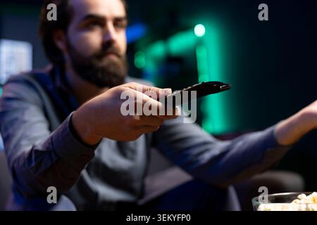 Tired man zapping through channels, watching news broadcast while resting in living room, enjoying leisure. Person using remote control to select TV station, close up shot Stock Photo