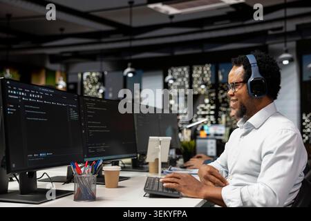 Cheerful technician wearing headphones doing code testing and quality assurance. Happy programmer in artificial intelligence focused startup designing and debugging applications, listening music Stock Photo