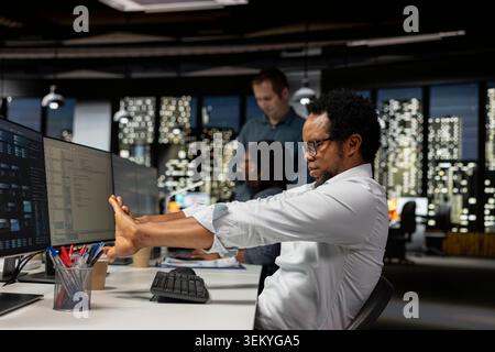 Tired programmer stretching joints in AI startup office, taking break to recover. Man at work doing body exercises at desk to relax muscles and eliminate fatigue from sitting on chair all day Stock Photo