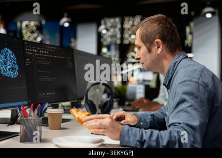 Admin eating croissant while using computer in tech startup office to develop programs with AI. IT professional enjoying snack break while debugging applications with artificial intelligence Stock Photo