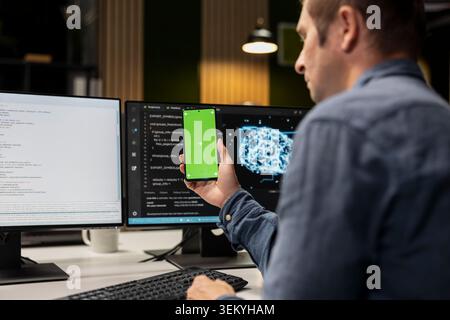 Green screen smartphone used by technician in startup office, optimizing code for performance and scalability. Mockup chroma key phone used by IT worker looking at programming language on PC Stock Photo