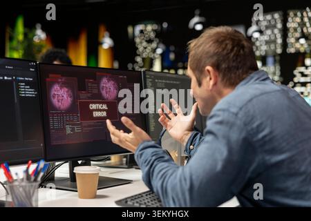Engineer at office desk frustrated by critical error warning displayed on computer screen. Upset sech startup IT professional annoyed by computer bug affecting server network Stock Photo