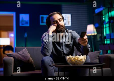 Cord cutter bored at home using TV to watch films on streaming services, zapping through content. Man in living room mindlessly consuming entertainment, using remote control Stock Photo