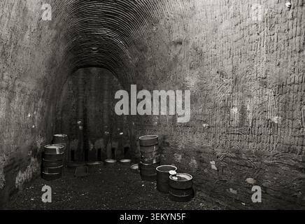 Vintage monochrome shot of an arched earthen pottery kiln interior, stacked cylindrical ceramic pots scattered across gritty dirt ground, rough textur Stock Photo