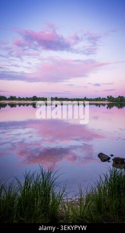 Pastel sunset over Obolon in Kyiv, Ukraine. Pink clouds reflect on calm water, creating a serene landscape with soft evening colors, shoreline grass, Stock Photo