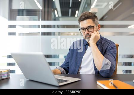 Man with glasses intently working on a laptop at a modern office desk, focused on problem solving and planning, embodying remote-friendly professional concentration and innovation Stock Photo