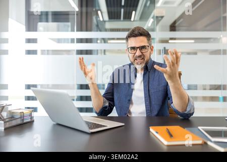 Stressed male employee experiencing a work problem, feeling overwhelmed, and expressing frustration with open hands while sitting at a modern office desk with a laptop Stock Photo