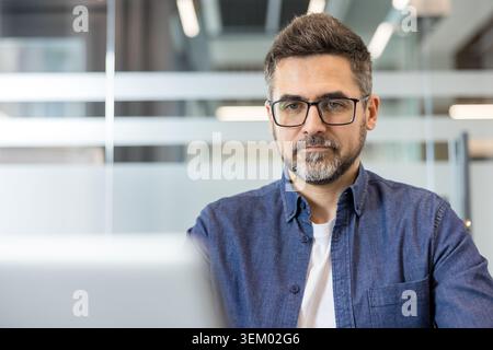 Professional bearded man wearing glasses focusing on his work, sitting at a laptop in a modern business office, representing dedication and productivity in a corporate environment Stock Photo