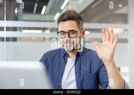 Man in office setting greeting on a laptop screen, participating in a remote video conference meeting using modern technology for virtual communication and working from home Stock Photo