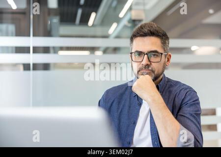 Serious bearded businessman with glasses gazes at camera in modern office, pensive and focused on strategy, planning, and future success for his startup or corporate team Stock Photo