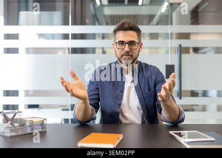 Mature business professional leading a meeting or presentation, gesturing with hands while communicating ideas and giving an online explanation in a modern office environment Stock Photo