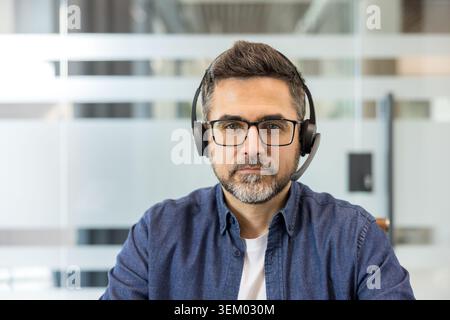Serious male call center agent or customer service representative looking directly at camera, wearing a headset with a microphone in a modern office environment Stock Photo