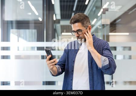 Mature man with glasses standing in a modern office, worried while checking his smartphone, touching his face after reading a troubling message or notification on screen Stock Photo