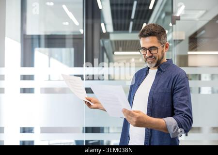 Confident man reviewing corporate paperwork in a contemporary office setting, focusing on data analysis, planning, and achieving business goals with a positive expression Stock Photo
