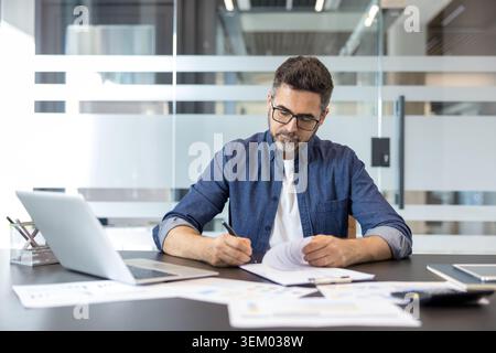 Businessman writing on a paper document while reviewing reports and financial data at his desk, demonstrating focus and professional engagement in a modern office environment Stock Photo