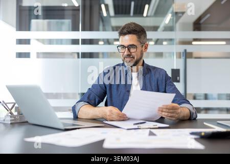 Confident businessman in glasses and denim shirt reads documents at a modern office desk, focused on laptop and paperwork while planning finances and strategy for his startup Stock Photo