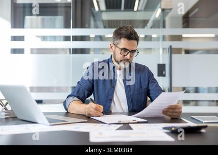Businessman wearing glasses and a denim shirt concentrating on signing a contract and reviewing financial reports at the modern office desk with a laptop Stock Photo