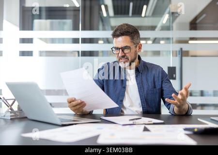 Senior businessman sitting at office desk, receiving bad news, looking stressed and frustrated while analyzing important documents and calculations with a laptop Stock Photo