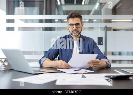 Confident male manager sitting at a desk in a contemporary office, carefully reviewing business documents and financial reports while looking directly at the camera Stock Photo