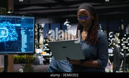 Female technician in office using laptop, working to fix bugs in artificial intelligence system. Woman troubleshooting neural network errors with notebook in tech agency workplace Stock Photo
