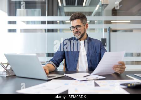 Businessman working at a modern office desk, reviewing financial documents and using a laptop, representing business efficiency and professional productivity Stock Photo