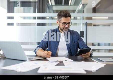 Businessman sitting at an office desk, evaluating financial reports with a calculator and writing notes, actively managing business finances and planning strategy Stock Photo