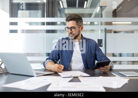 Mature bearded businessman working at an office desk, writing on documents, and using a calculator while a laptop and financial reports are spread out Stock Photo
