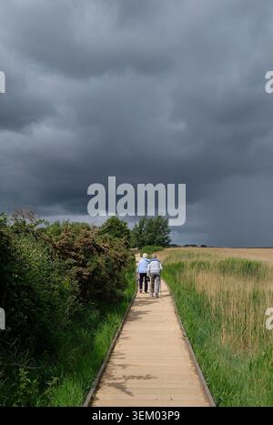 elderly couple walking towards storm clouds on boardwalk, brancaster, north norfolk, england Stock Photo